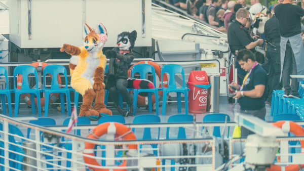 Two fursuiters sit on the upper deck of the boat whilst others wait to board